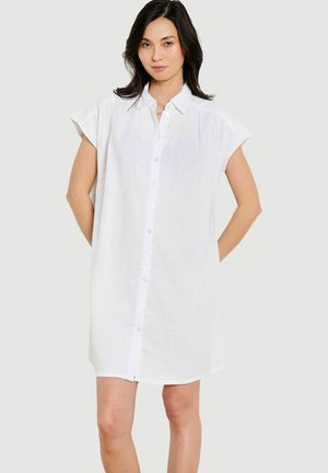 Femme portant une robe chemise blanche sans manches avec col, debout devant un fond clair uni, mains dans le dos.