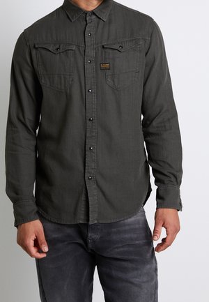 Homme portant une chemise à manches longues gris foncé boutonnée avec deux poches poitrine et un jean noir délavé, debout devant un fond uni.