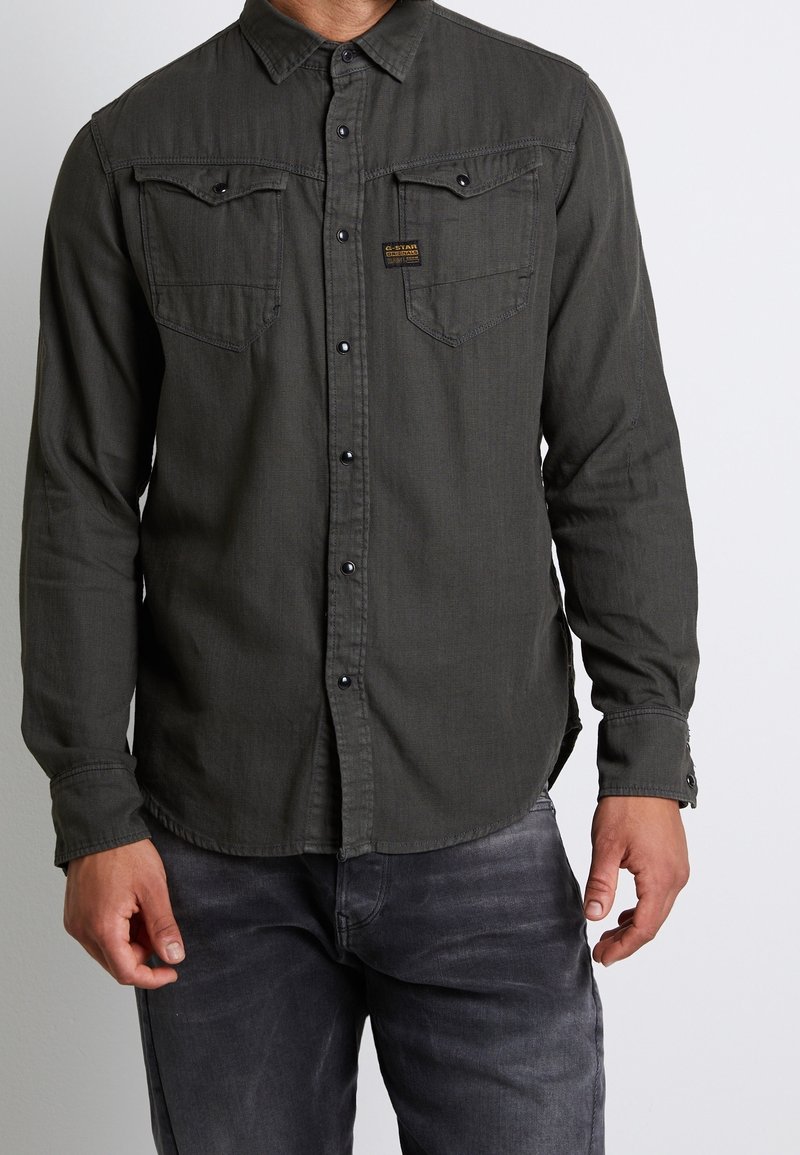 Homme portant une chemise à manches longues gris foncé boutonnée avec deux poches poitrine et un jean noir délavé, debout devant un fond uni.