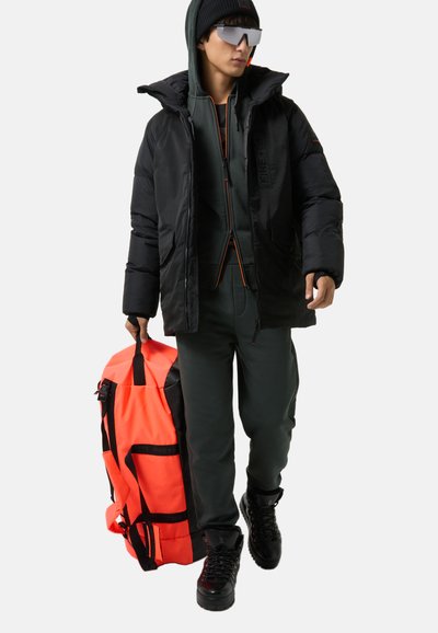 Black puffer jacket with orange accents, green tracksuit, and black boots. Holding a large, bright orange and black duffle bag.