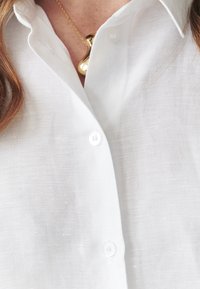 Camicia bianca in lino con chiusura a bottoni e colletto rigido, abbinata a una collana con pendente dorato. È visibile una trama tessile.