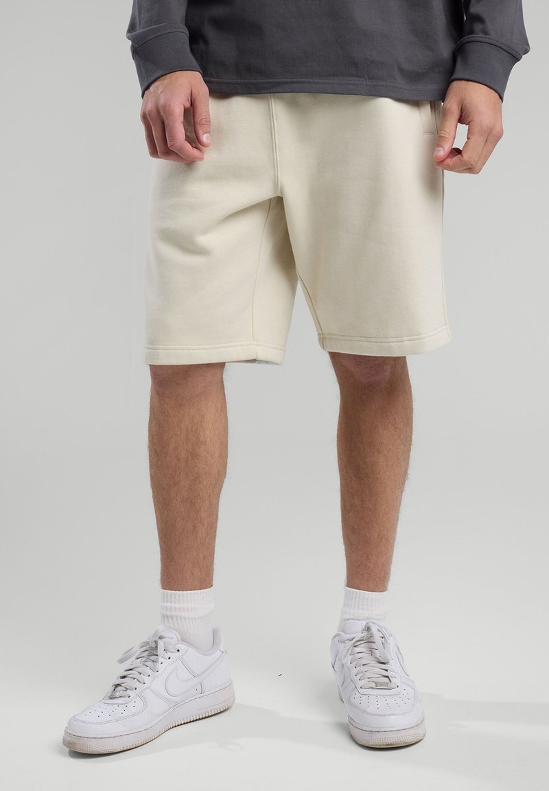 Kermanväriset, keskipituiset shortsit, jotka on valmistettu pehmeästä kankaasta. Shortsissa on sivutaskut ja näkyvät ompeluyksityiskohdat, ja ne on yhdistetty valkoisiin urheilukengiin ja sukkiin.