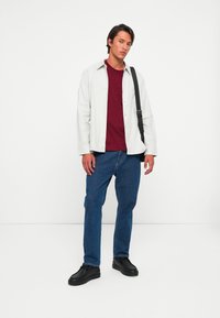 Tânăr stând cu o geantă neagră pe umăr, purtând o cămașă albă deschisă peste un tricou roșu, blugi și pantofi negri, pe un fundal alb simplu.