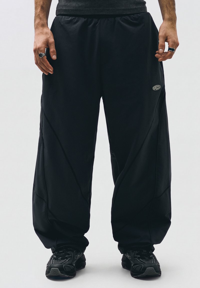 Homme portant un pantalon noir ample, des baskets noires et des bagues aux deux mains, debout devant un fond clair uni.