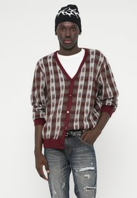 Καρντιγκάν σε μπορντό, γκρι και κρεμ καρώ σχέδιο, με V-neck, μπροστινές κουμπί και  ribbed μανσέτες. Φορεμένο πάνω από λευκή μπλούζα, συνδυασμένο με τζιν με φθορές.