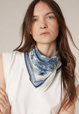 Femme aux longs cheveux bruns portant un haut blanc sans manches et une écharpe à motifs bleu et blanc autour du cou.