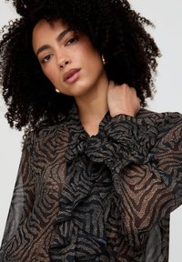 Transparente schwarze Bluse mit wirbelndem braunem Muster, mit Rüschen am Kragen und den Ärmeln sowie strukturiertem Stoff.