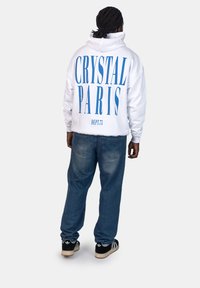 Felpa bianca con grande testo blu "CRYSTAL PARIS" sulla schiena, abbinata a jeans blu e sneakers nere. Il tessuto appare morbido con una vestibilità rilassata.