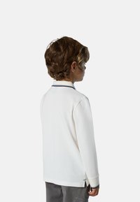 Camisa polo blanca de manga larga con un acento azul marino en el collar, de tela texturada y con aberturas laterales, usada por un niño.