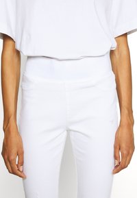 Pantalones de mezcla de algodón blancos con una alta cintura, que cuentan con bolsillos frontales y una superficie lisa y ligeramente texturizada. Diseño sencillo y minimalista.
