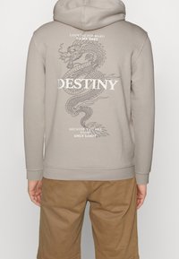 Sudadera beige con un gráfico de dragón en la parte posterior. El texto incluye "DESTINO" y frases motivacionales. Material suave y puños acanalados.