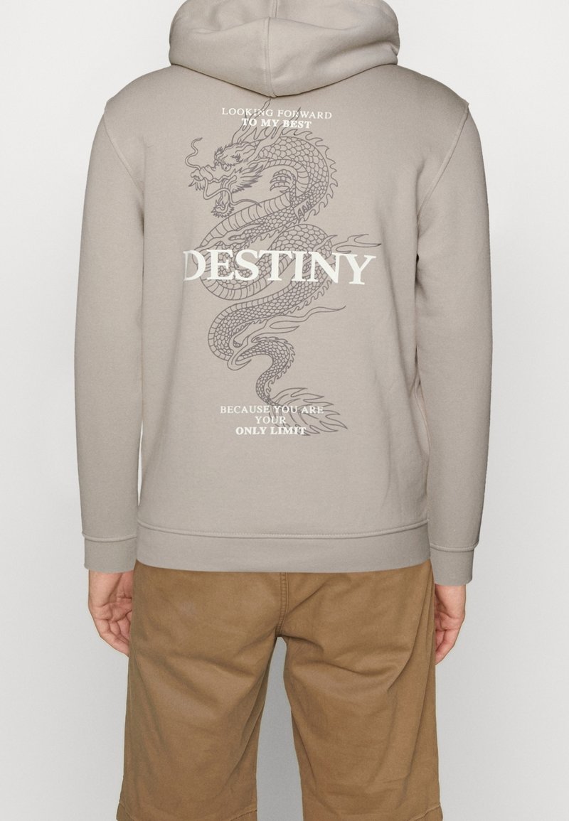 Sudadera beige con un gráfico de dragón en la parte posterior. El texto incluye "DESTINO" y frases motivacionales. Material suave y puños acanalados.