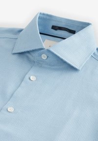 Chemise bleu clair à manches longues avec un motif texturé, dotée d'un col évasé et de boutons blancs sur le devant.