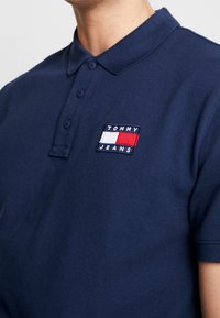 Námořnická modrá polo košile z texturované látky s dvouknoflíkovou légou. Obsahuje vyšitý obdélníkový logo s textem "TOMMY" a "JEANS".