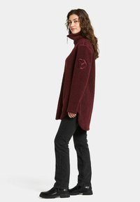 Burgunderröd fleecetröja med hög krage och sidofickor, med en texturerad yta och en logodetalj på ärmen. Bärs med svarta jeans.