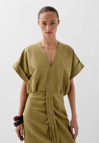 Olijfgroene jurk met een v-hals, korte mouwen en ruches op de taille. Het model draagt donkere armbanden aan één pols.