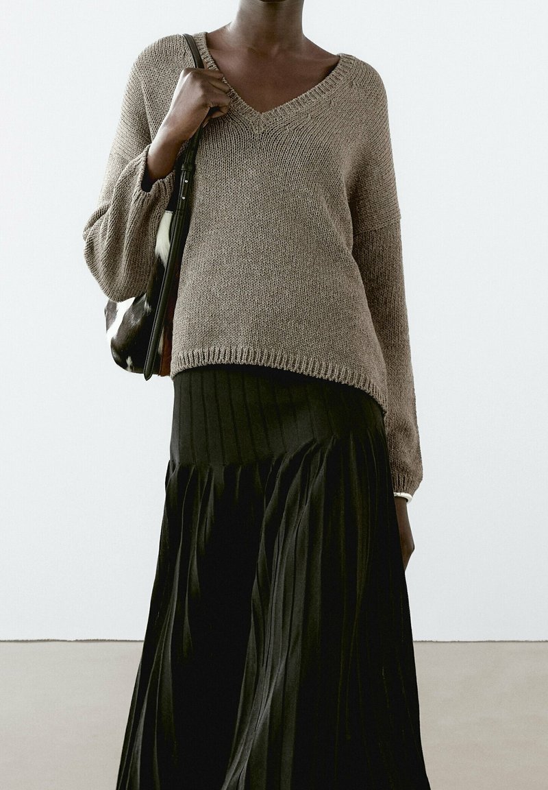 Pull en maille marron à col en V avec des manches larges, associé à une jupe noire plissée et fluide. La tenue est complétée par un sac à bandoulière au motif distinctif.