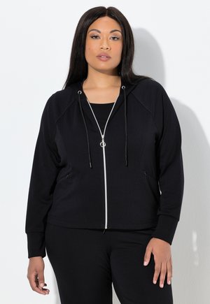 Sweat à zip noir avec fermeture éclair argentée, cordon de serrage et poches latérales. Tissu doux, manches raglan, associé à un haut et un pantalon noirs.