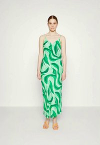 Vestido maxi com padrão verde apresentando designs abstratos semelhantes a folhas, com alças finas e uma silhueta fluida, combinado com sandálias laranja com tiras.