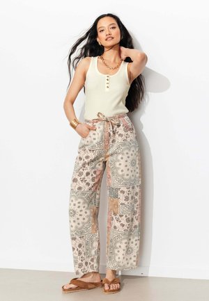 Femme portant un haut sans manches crème, un pantalon large à motifs avec une taille à cordon, des bijoux en or et des sandales marron, posant contre un mur blanc.