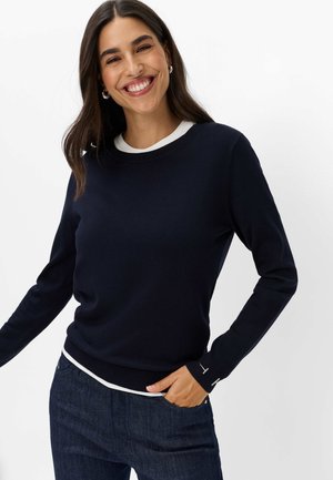 Vrouw glimlacht en draagt een marineblauwe trui met lange mouwen en witte bies, en donkerblauwe jeans, staande voor een effen achtergrond.
