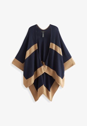 Kvinde går iført en marineblå og tan åben poncho, sort top med blondetrim, mørke jeans, sorte ankelstøvler og bærer en brun crossbody-taske.