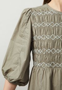 Sukienka z bawełny w kolorze oliwkowym z marszczonym ciałem, bufiastymi rękawami i białymi haftowanymi detalami. Posiada marszczony dół i szwy boczne.