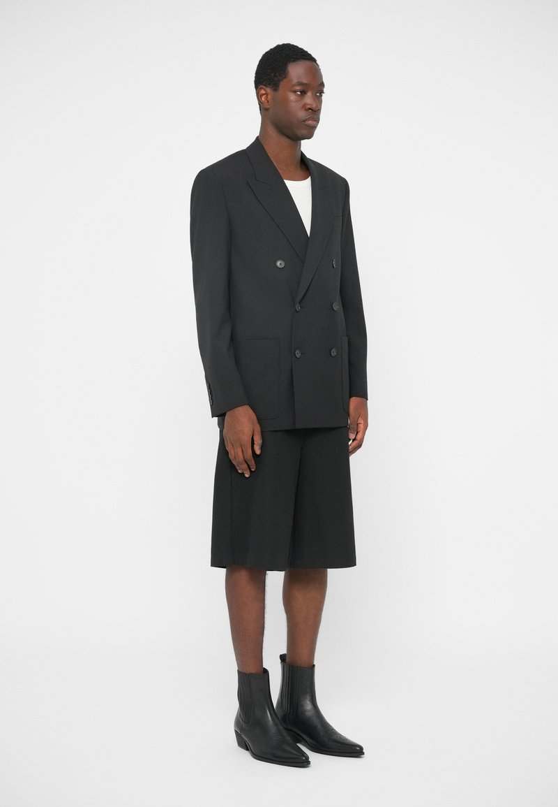 Sort dobbeltknappet blazer i en struktureret design, parret med knælange sorte shorts og sorte ankelstøvler, sat mod en hvid baggrund.