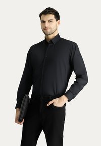 Camisa negra de manga larga con botones y cuello estructurado. La tela es suave con un ligero brillo. Combinada con jeans negros y un bolso de mano texturizado.