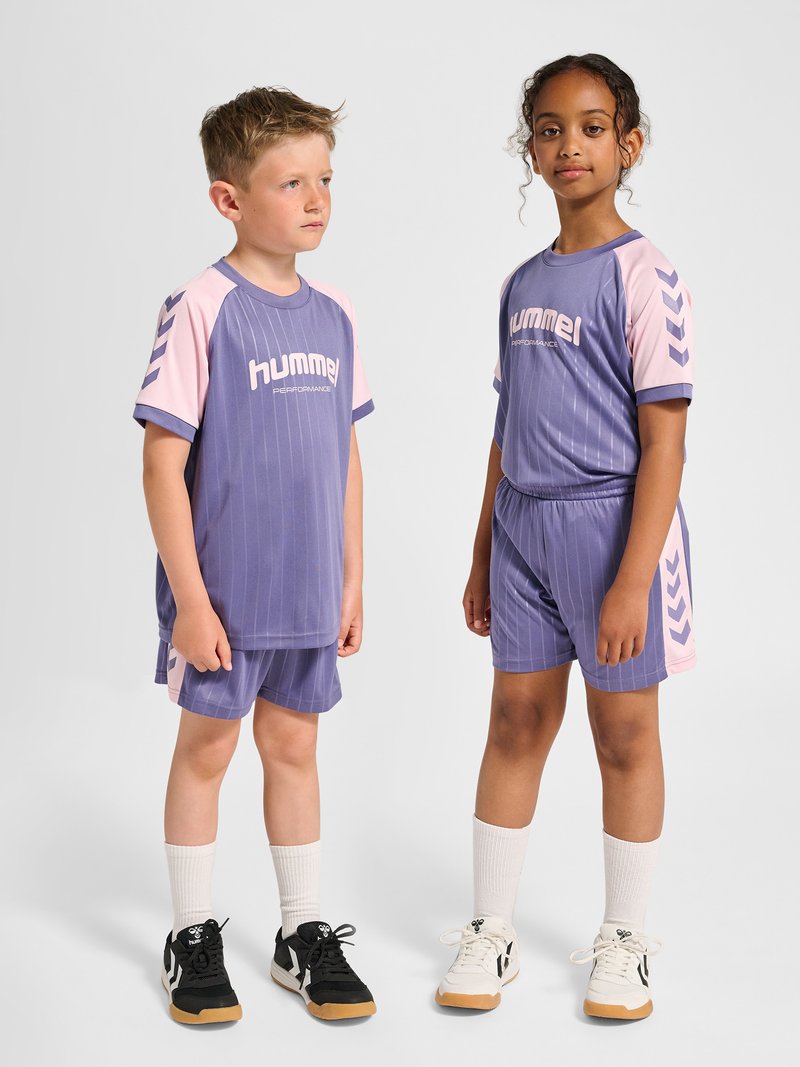 Zwei Kinder stehen, tragen passende lila und pinke Hummel-Sportkleidung mit weißen Socken und Turnschuhen, vor einem einfarbigen Hintergrund.
