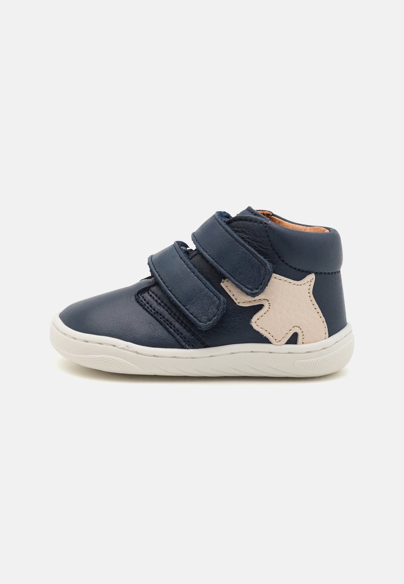 Chaussure haute en cuir bleu marine avec deux bandes Velcro, semelle blanche et étoile beige texturée sur le côté. Conçue pour le confort et le soutien.