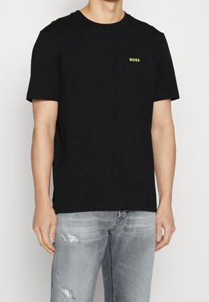 Homme portant un t-shirt noir uni à manches courtes avec un petit texte jaune-vert "BOSS" sur la poitrine gauche et un jean gris clair usé.