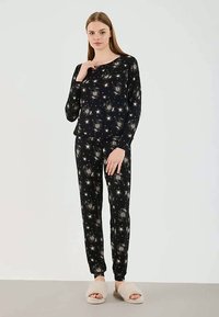 Woman standing wearing black long-sleeve top and pants with white star patterns, and beige fuzzy slippers on a plain background.