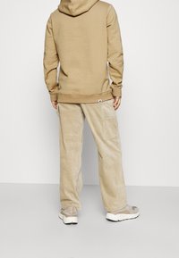 Felpa beige con cappuccio e orlo a coste, abbinata a pantaloni in velluto a coste beige con tasche laterali. Il modello indossa sneakers beige.