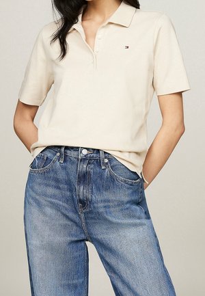 Kvinde iført off-white kortærmet polo skjorte med lille logo på brystet, stoppet ned i blå høj talje jeans mod neutral baggrund.