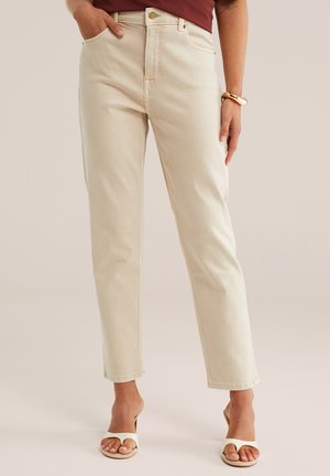 Kobieta w beżowych dżinsach z wysokim stanem i prostymi nogawkami, bordowej bluzce, beżowych sandałkach na obcasie z odkrytymi palcami oraz beżowej bransoletce.