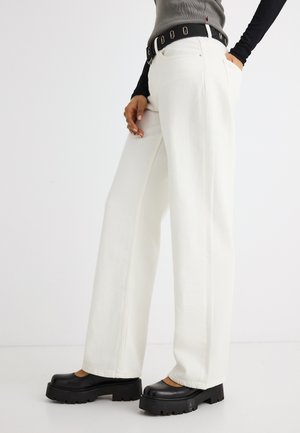 Személy széles szárú fehér nadrágot, fekete vastag talpú cipőket, szürke felsőt fekete hosszú ujjakkal és ezüst szegecsekkel díszített fekete övet visel.