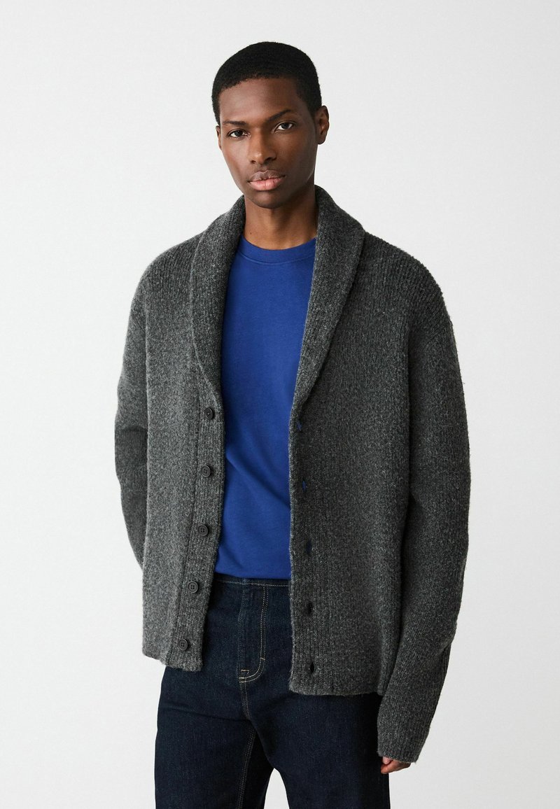 Dark gray knitted cardigan with a shawl collar, front button closure, and textured fabric. Paired with a blue shirt and dark jeans.