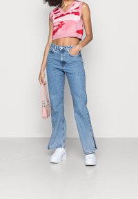 Haut en tricot rose coupé avec des motifs rouges abstraits, associé à un jean évasé bleu clair avec des fentes latérales, et des baskets compensées blanches.