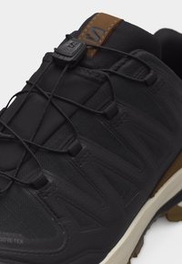 Μαύρο παπούτσι πεζοπορίας με σύστημα ταχείας δέσης, καφέ λουράκι στη φτέρνα και λευκή σόλα, με υφή συνθετικού υλικού και υφασμάτινη επάνω πλευρά.