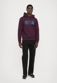 Σκούρο μοβ φούτερ με κουκούλα και ανοιχτό μπλε λογότυπο "VERSACE JEANS COUTURE", συνδυασμένο με μαύρο παντελόνι και μαύρα αθλητικά παπούτσια.