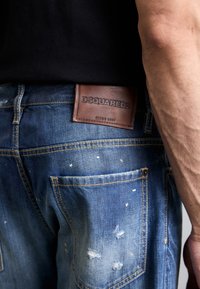 Ošuntělé modré džíny s lehkými skvrnami od barvy, s hnědou koženou nášivkou DSQUARED2 vzadu a žlutými švy.