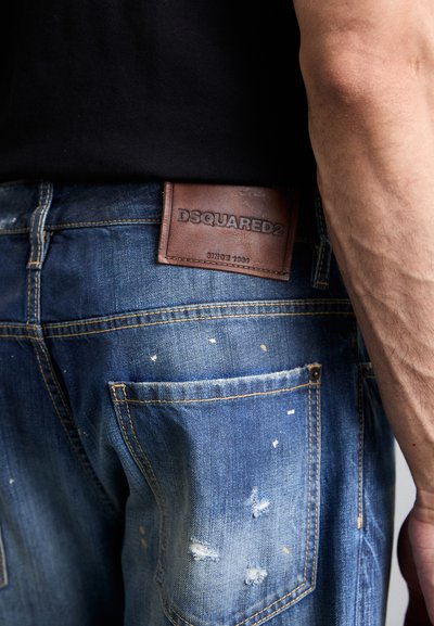 Kulot denim biru yang distressed dengan percikan cat terang, dilengkapi label kulit DSQUARED2 berwarna coklat di bagian belakang dan detail jahitan kuning.