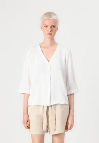 White button-up shirt with a V-neck and 3/4 sleeves, made from lightweight fabric. Paired with beige linen shorts.