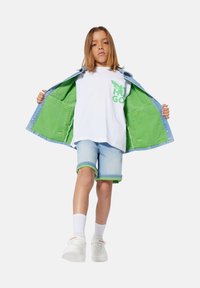 Chaqueta de mezclilla con forro verde, camiseta blanca con estampado verde, pantalones cortos azul claro con puños verdes y zapatillas blancas con logo.