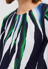 Detailní záběr osoby v blůze s kulatým výstřihem, která má abstraktní námořnické, zelené, modré a bílé vlnité pruhy.