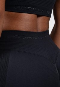 Black athletic leggings feature a textured waistband with tonal branding, smooth fabric, and distinct panel detailing for breathability.
