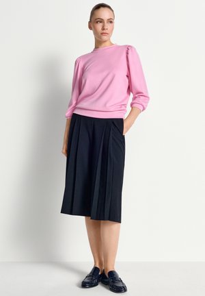 Nő áll, kezét a zsebében tartja, rózsaszín hosszú ujjú felsőt, fekete pliszírozott szoknyát és fekete loafert visel, fehér háttér előtt.