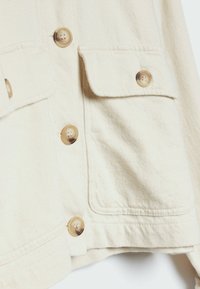 Kremowa kurtka z teksturowanej tkaniny, z dwiema klapkowymi kieszeniami, pięcioma guzikami i zaokrąglonymi guzikami o lekko błyszczącym wykończeniu.