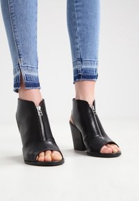 Svarta läderöppna klackstövlar med framdragkedja, blockklack och utskärningsdesign. Bärs med slitna blå skinnyjeans.